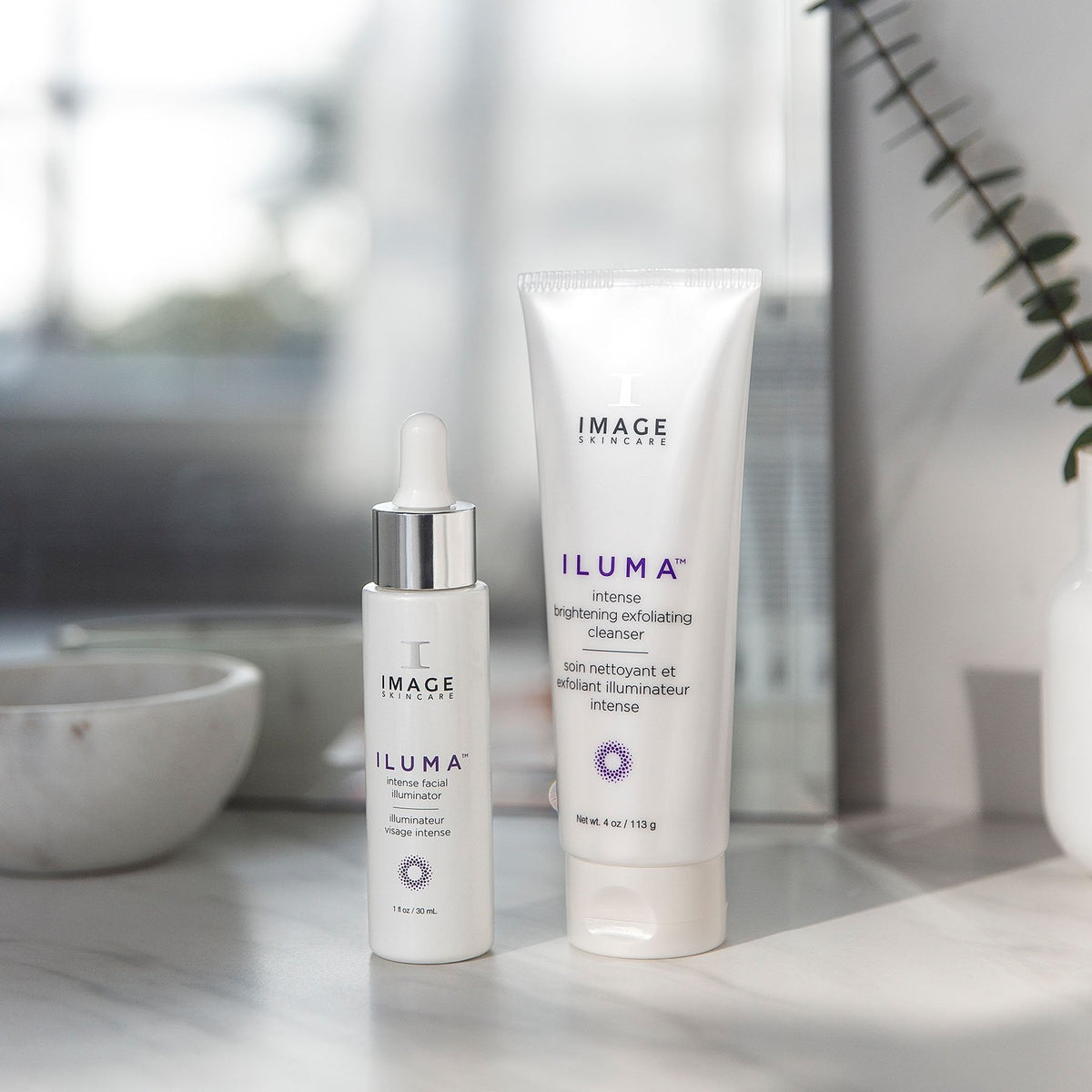IMAGE Skincare ILUMA Intense Brightening Exfoliating Cleanser with ILUMA Intense Facial Illuminator on a bathroom shelf
